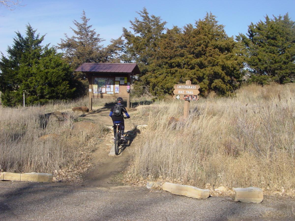 A person riding a mountain bike on a trail leading toward a trailhead sign for the Switchgrass Bike Trail, with a wooden information kiosk in the background surrounded by grass and shrubs. Switchgrass mountain bike trail.