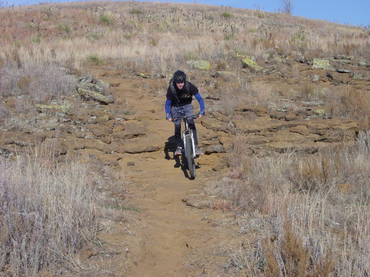 Mountain biker navigating a rocky trail with dry grass and shrubs in the background on a sunny day. Switchgrass mountain bike trail.