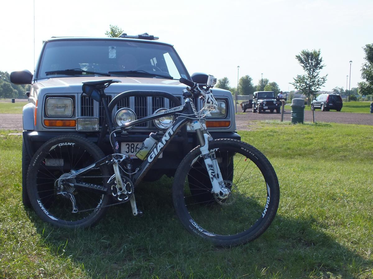 Giant Anthem 1: A mountain bike leaning against the front of a white Jeep in a grassy parking area, with additional vehicles parked in the background and trees lining the scene under a clear sky.