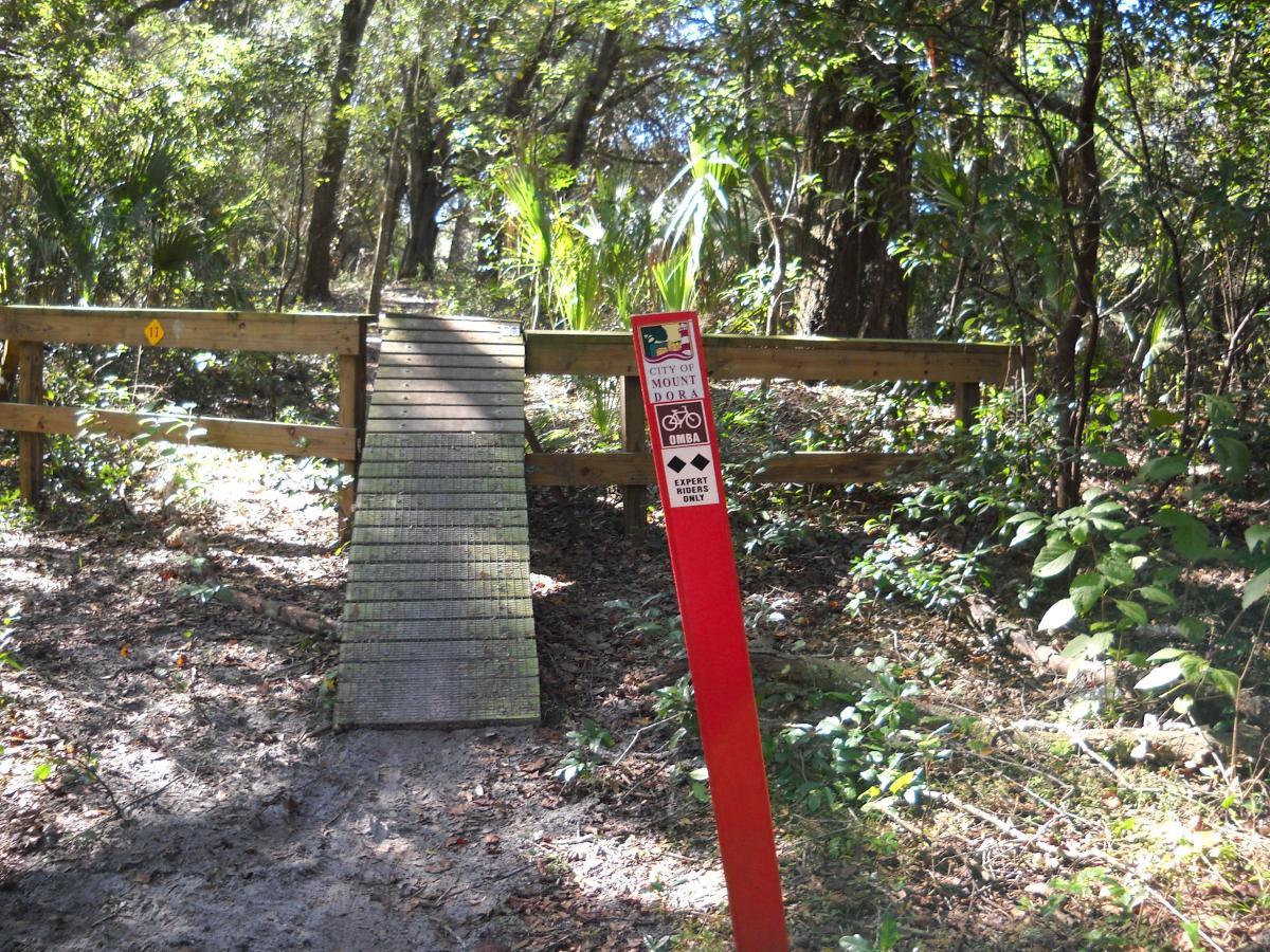 A wooden bridge leading into a wooded area, with signs indicating a mountain biking trail. The trail is marked for expert riders only. Lush greenery surrounds the bridge, with sunlight filtering through the trees. Mount Dora Trail mountain bike trail.