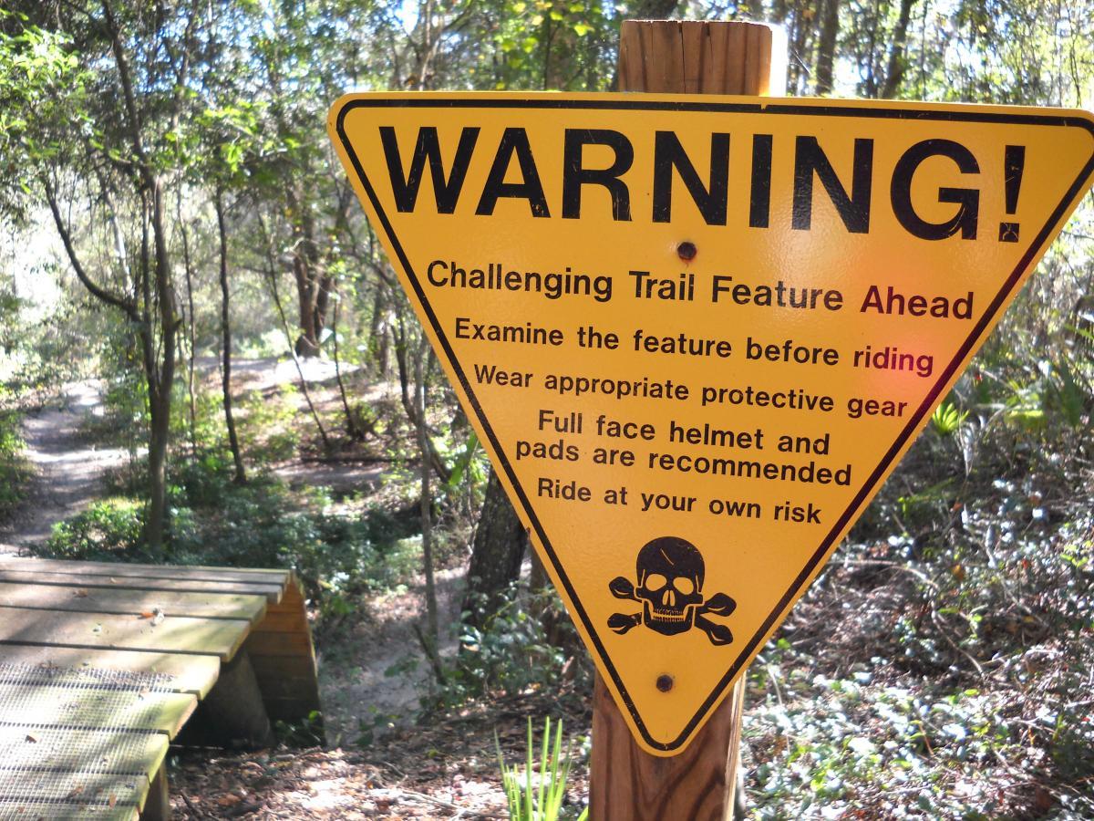 A warning sign at a trailhead indicating the presence of a challenging trail feature. The sign advises riders to examine the feature before riding, wear appropriate protective gear such as a full-face helmet and pads, and to ride at their own risk. The background features a wooded area with a dirt path leading into the trees. Mount Dora Trail mountain bike trail.