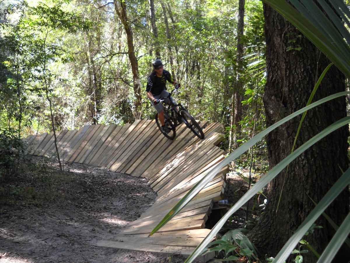 A mountain biker rides a wooden pump track through a forested area, leaning into a turn on the curved section of the track surrounded by lush greenery and trees. Mount Dora Trail mountain bike trail.