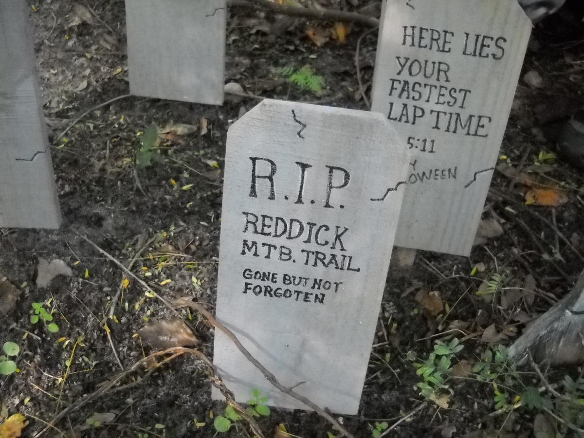 A humorous wooden gravestone in a wooded area, reading "R.I.P. Reddick MTB Trail, Gone But Not Forgotten." In the background, there is another gravestone that says "Here Lies Your Fastest Lap Time 5:11." The setting features fallen leaves and small plants around the gravestones. Mount Dora Trail mountain bike trail.