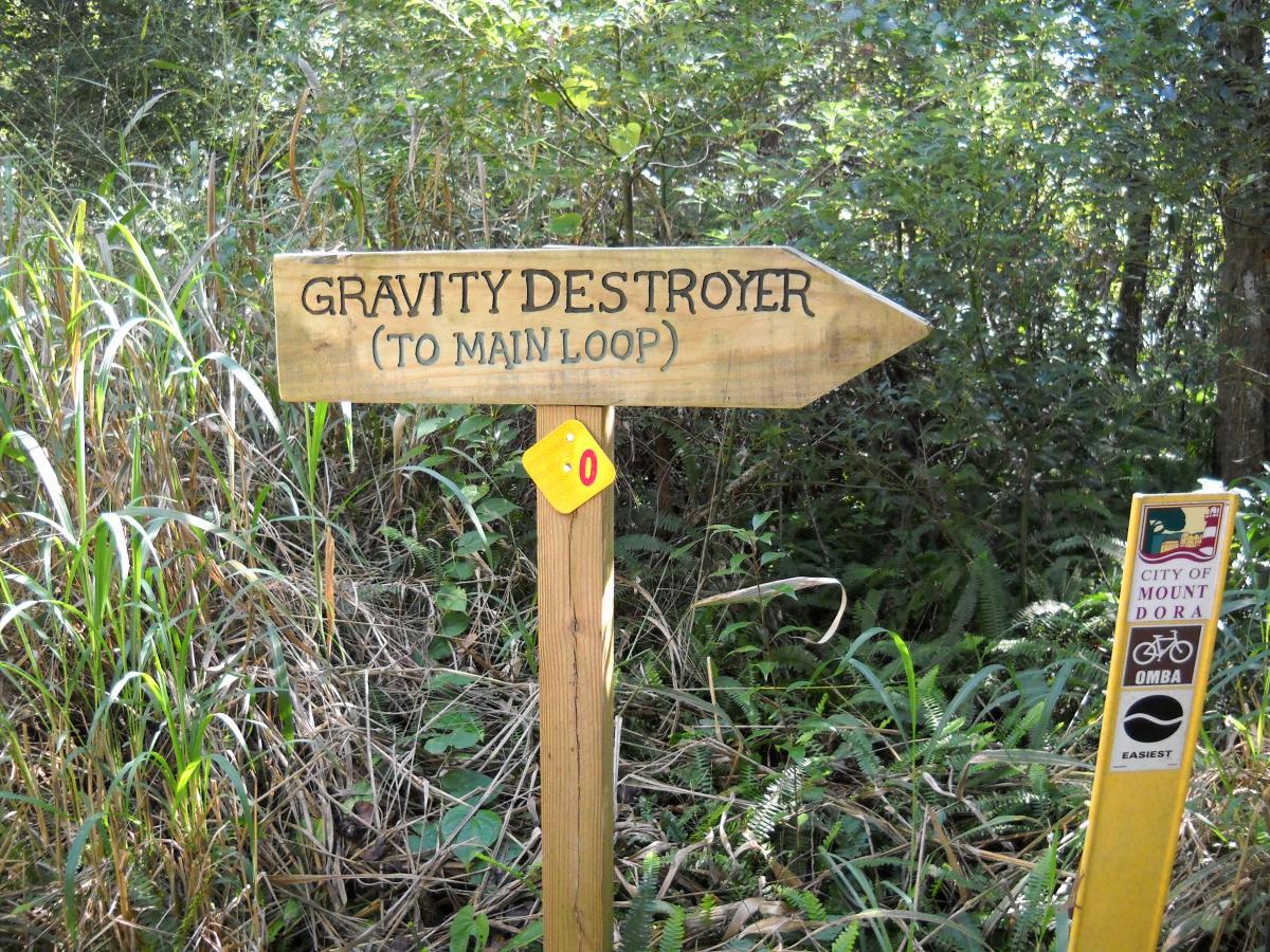 A wooden signpost in a forested area pointing left towards "Gravity Destroyer (to main loop)," with a yellow trail marker and additional informational signage on the right about mountain biking trails in the City of Mount Dora. Lush green foliage surrounds the pathway. Mount Dora Trail mountain bike trail.