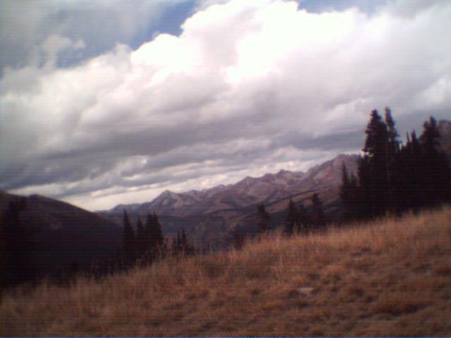 A scenic view of mountainous terrain under a cloudy sky. The foreground features dry grass and scattered trees, while the background showcases a range of rugged mountains with peaks under a dramatic cloudscape. Trail 401 mountain bike trail.