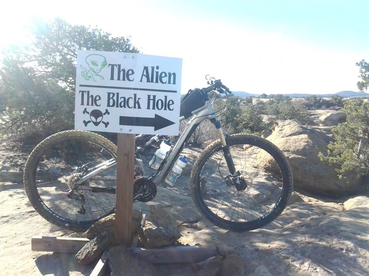 Trek Rumblefish Elite: Mountain bike parked next to a sign indicating directions to "The Alien" and "The Black Hole," featuring graphics of an alien and skulls, set against a rocky landscape and bright sunlight.
