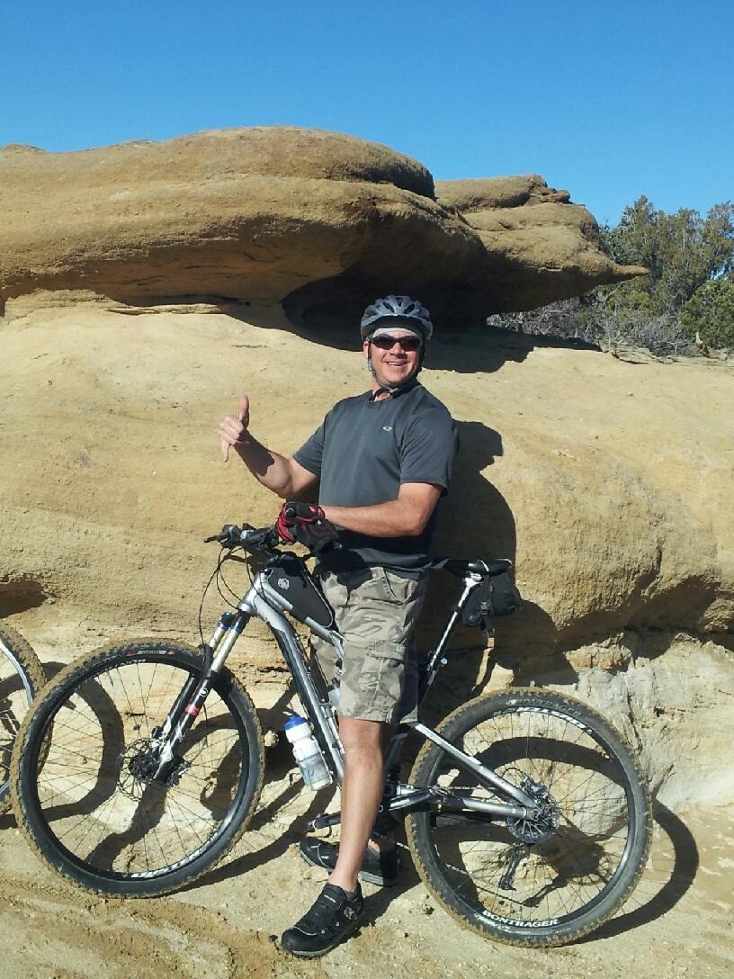 Trek Rumblefish Elite: A man stands beside his mountain bike, giving a thumbs-up gesture. He is wearing a helmet, sunglasses, and casual athletic clothing, with the backdrop featuring a large rock formation and a clear blue sky. The terrain is sandy and rocky, indicating an outdoor cycling environment.
