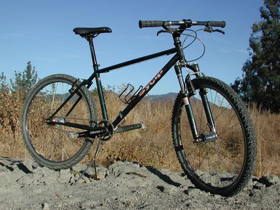 Sycip Diesel Single Speed: A black mountain bike with a sturdy frame and wide tires, positioned on a rocky surface with a background of dry grass and scattered trees under a clear blue sky.