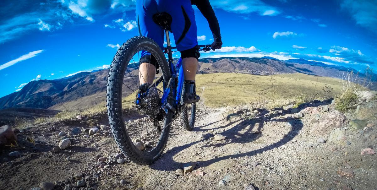 A cyclist riding a mountain bike on a rocky trail, with a scenic view of rolling hills and a clear blue sky in the background. The bike features thick tires, and the cyclist is dressed in blue athletic gear. Green Mountain mountain bike trail.
