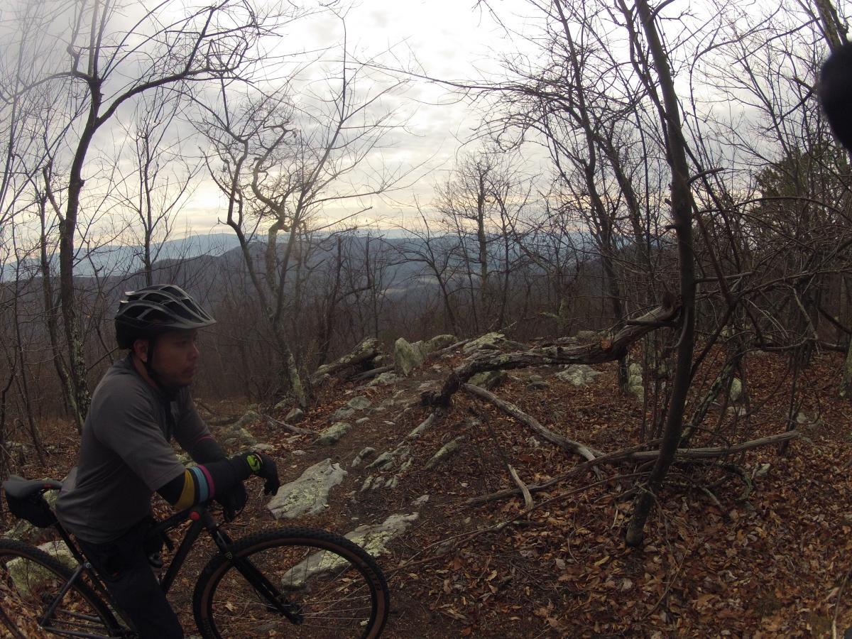 A cyclist wearing a helmet and gloves stands next to a mountain bike on a rocky trail surrounded by bare trees and autumn leaves. In the background, a mountainous landscape is visible under a cloudy sky. Elizabeth Furnace mountain bike trail.