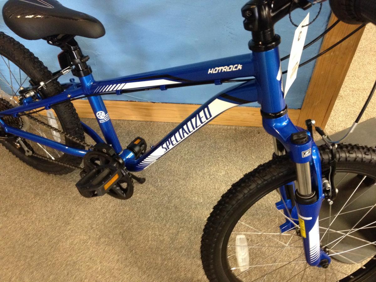 A blue Specialized Hotrock bicycle, showcasing its sturdy frame, black pedals, and thick tires, displayed in a shop setting with light-colored flooring and a blue wall in the background.