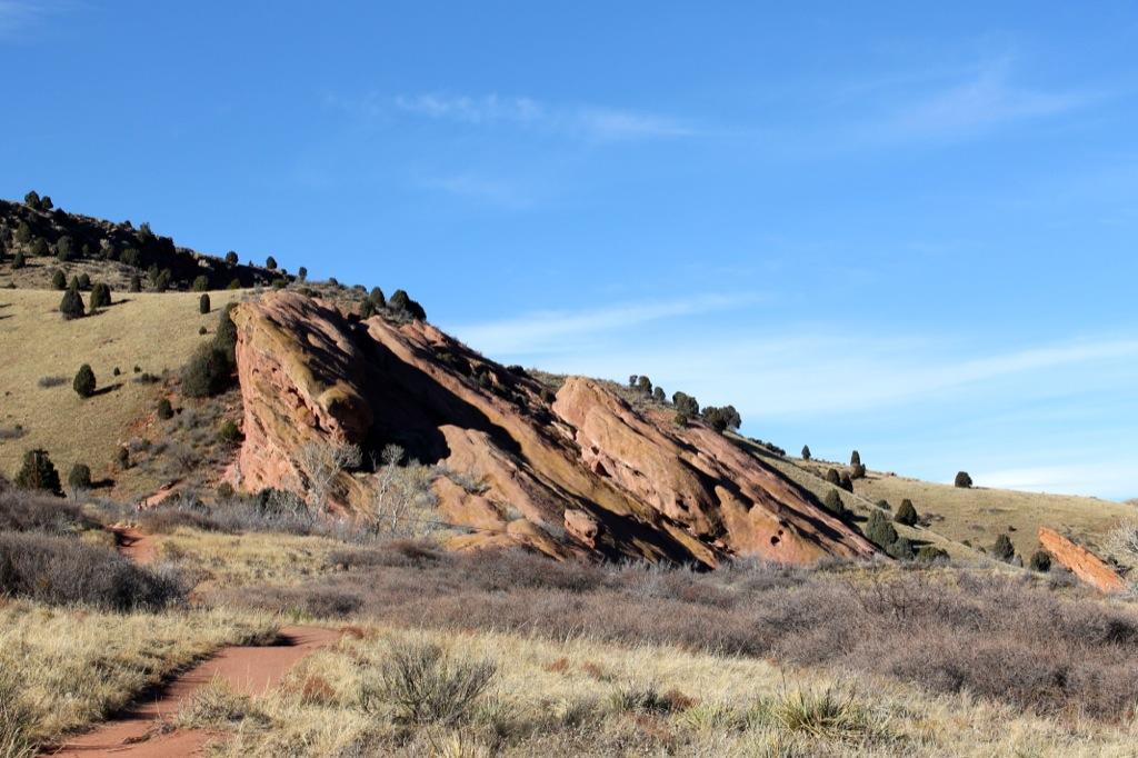 A scenic view of a rocky landscape featuring a prominent red rock formation on a hillside, surrounded by sparse vegetation and evergreen trees under a clear blue sky. A dirt path winds through the foreground, leading towards the rocks. Red Rocks / Dakota Ridge mountain bike trail.