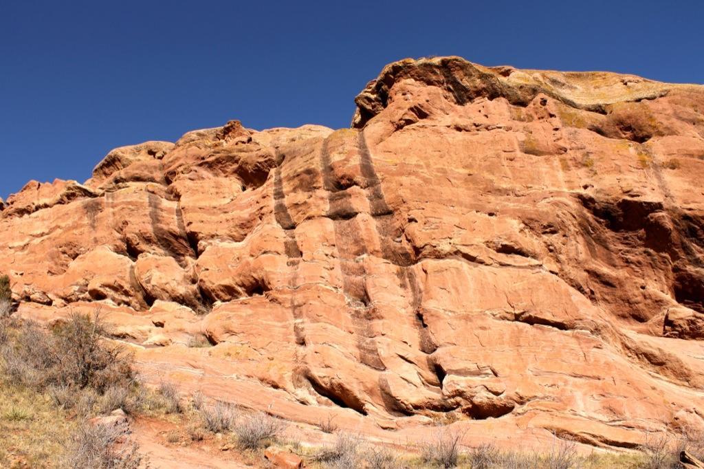Red rock formations with distinct, natural lines and textures against a clear blue sky. Sparse vegetation can be seen in the foreground. Red Rocks / Dakota Ridge mountain bike trail.