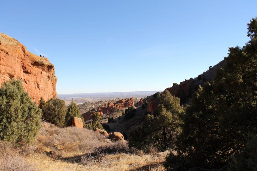 A scenic landscape featuring red rocky cliffs and greenery, with a clear blue sky. The view captures the vastness of the surrounding area, highlighting a natural valley in the distance. Red Rocks / Dakota Ridge mountain bike trail.