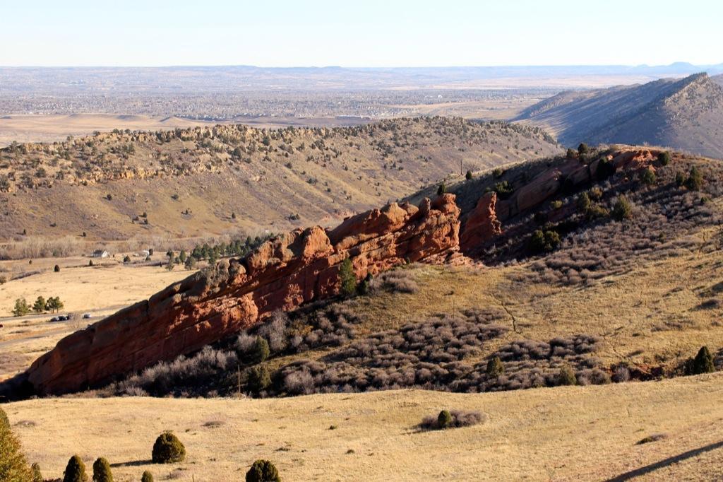 A scenic view of a rugged landscape featuring a prominent red rock formation on a sloped hillside, surrounded by rolling hills and sparse vegetation. The background showcases distant mountains and a clear blue sky. Red Rocks / Dakota Ridge mountain bike trail.