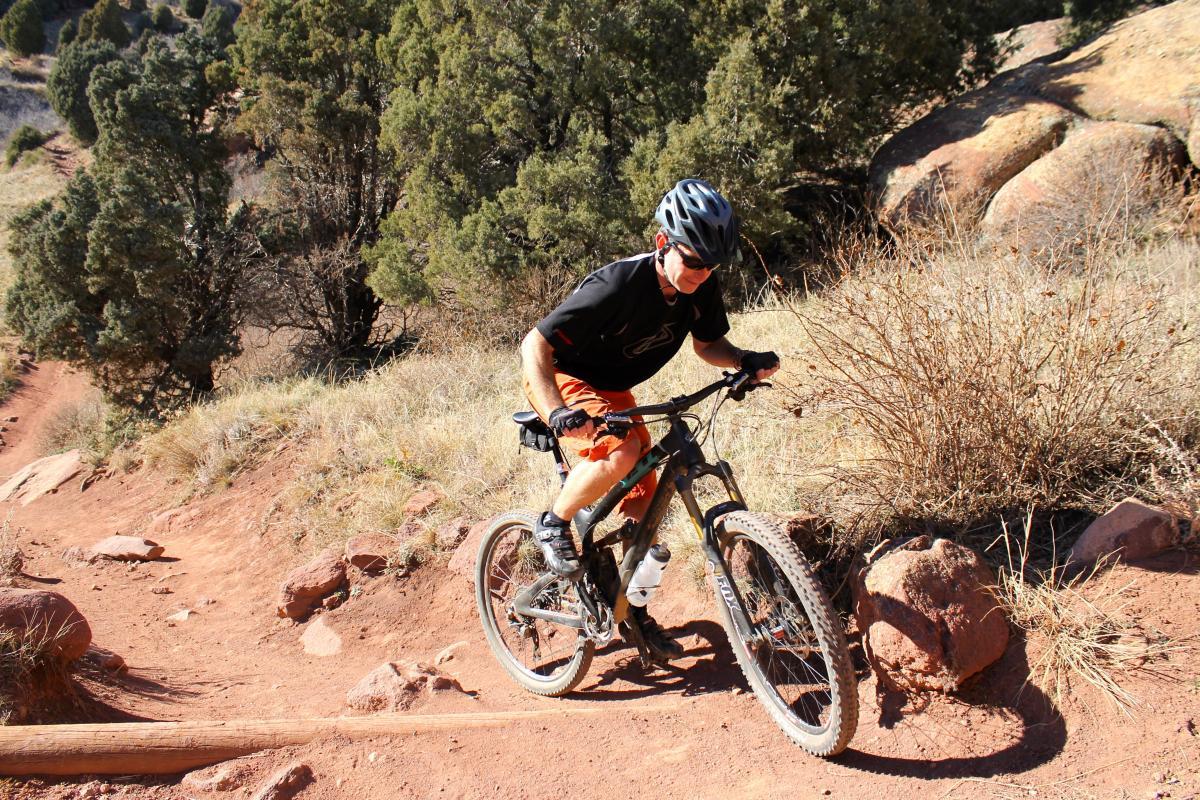 A person riding a mountain bike on a dirt trail, navigating a rocky section surrounded by trees and dry grass. The cyclist is dressed in a black shirt and orange shorts, wearing a helmet and gloves for safety. Red Rocks / Dakota Ridge mountain bike trail.