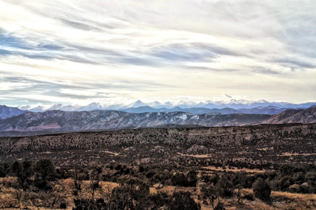 A panoramic view of a mountainous landscape featuring rugged peaks in the distance, partially covered in snow, under a sky filled with soft, wispy clouds. The foreground consists of rolling hills and sparse vegetation, creating a serene natural setting. Oil Well Flats mountain bike trail.