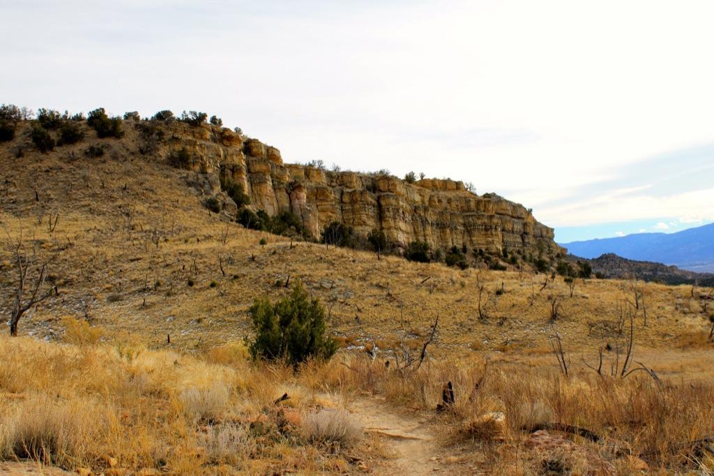 A rugged landscape featuring a rocky cliffside on the left, with dry grass and sparse vegetation in the foreground. A narrow dirt path winds through the scene, leading toward the foothills in the distance under a cloudy sky. Oil Well Flats mountain bike trail.