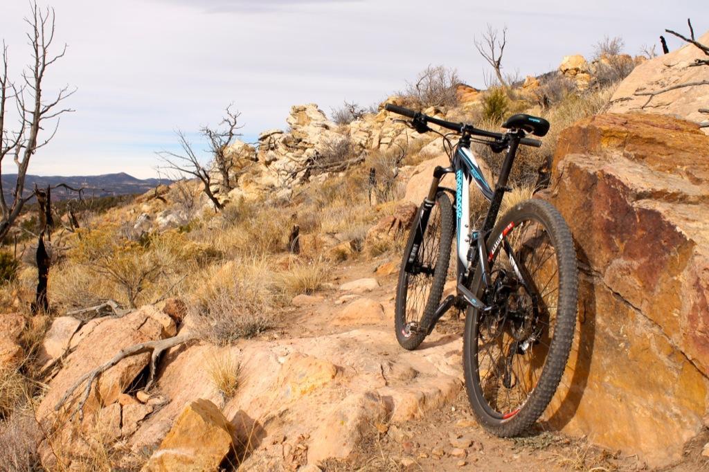 A mountain bike leaning against a large rock on a rugged trail surrounded by dry grass and sparse trees, with a distant mountainous landscape under a cloudy sky. Oil Well Flats mountain bike trail.