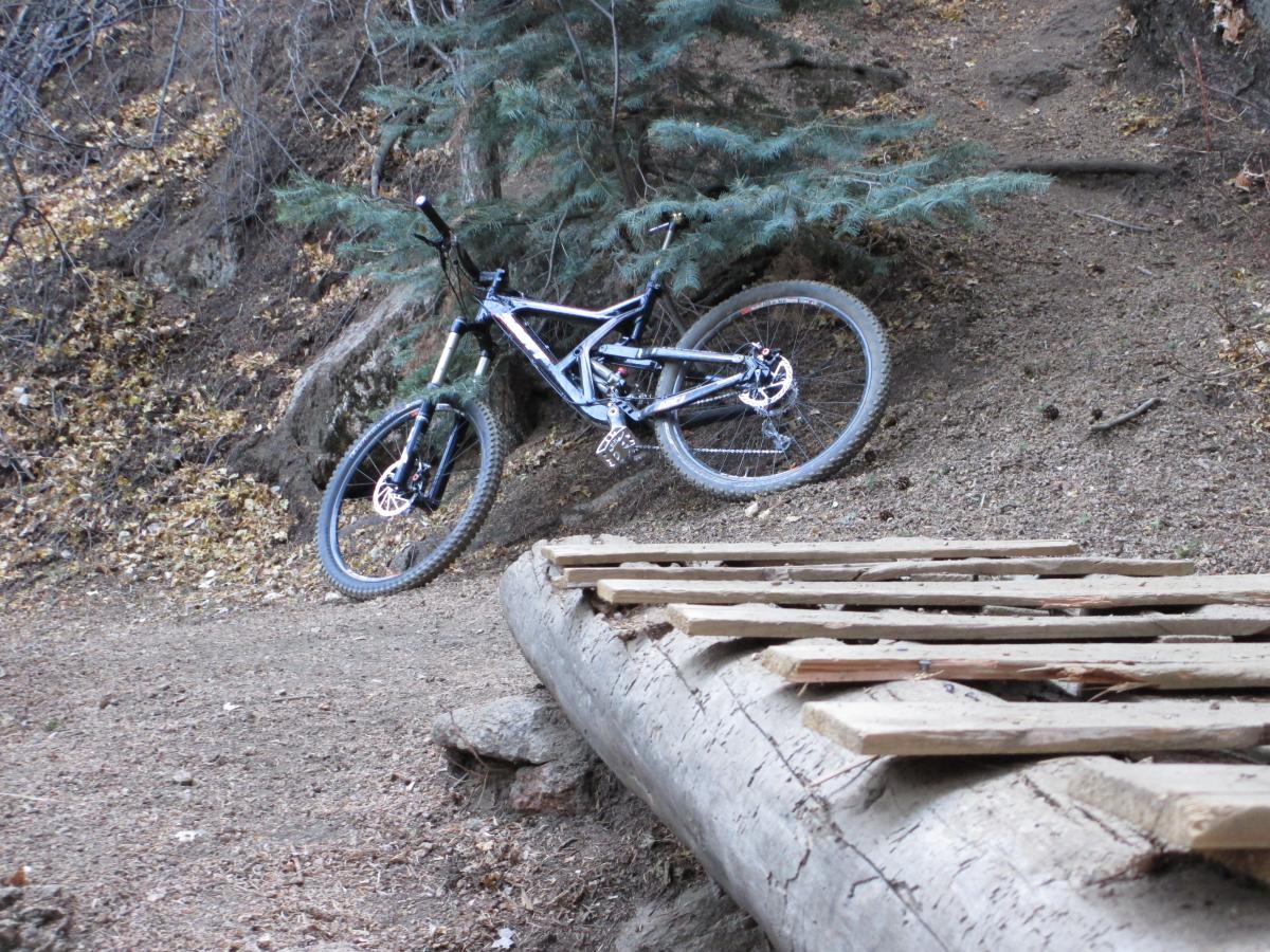 A mountain bike resting beside a wooden bridge on a dirt trail, surrounded by trees and fallen leaves. The terrain is uneven, indicating a natural outdoor setting suitable for biking. Palmer Trail / Section 16 mountain bike trail.