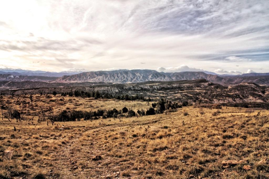 A panoramic view of rolling hills and mountain ranges under a cloudy sky, with dry, grassy terrain in the foreground and sparse trees scattered throughout. The landscape conveys a sense of openness and natural beauty. Oil Well Flats mountain bike trail.