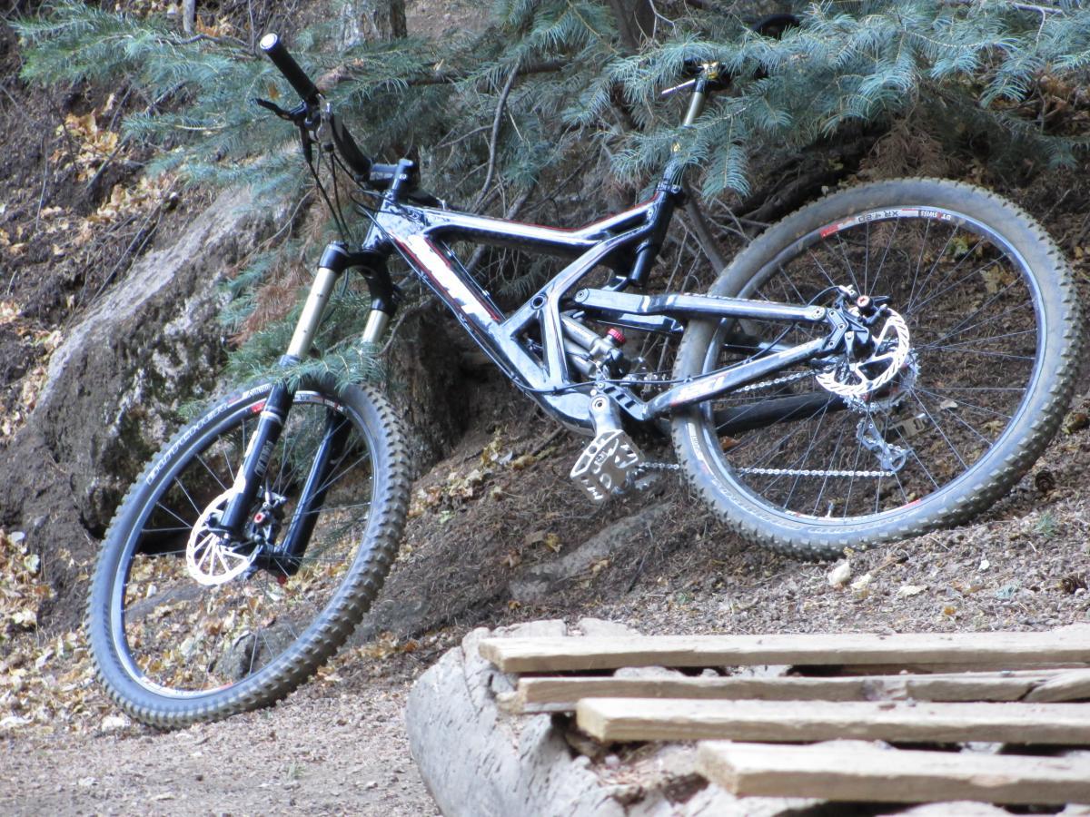 Scott Ransom: A mountain bike resting on a rocky slope, partially obscured by trees. The bike features a sturdy frame and knobby tires, positioned at an angle near a wooden structure on the ground. The surrounding environment includes earthy tones and scattered foliage, indicating a natural outdoor setting.