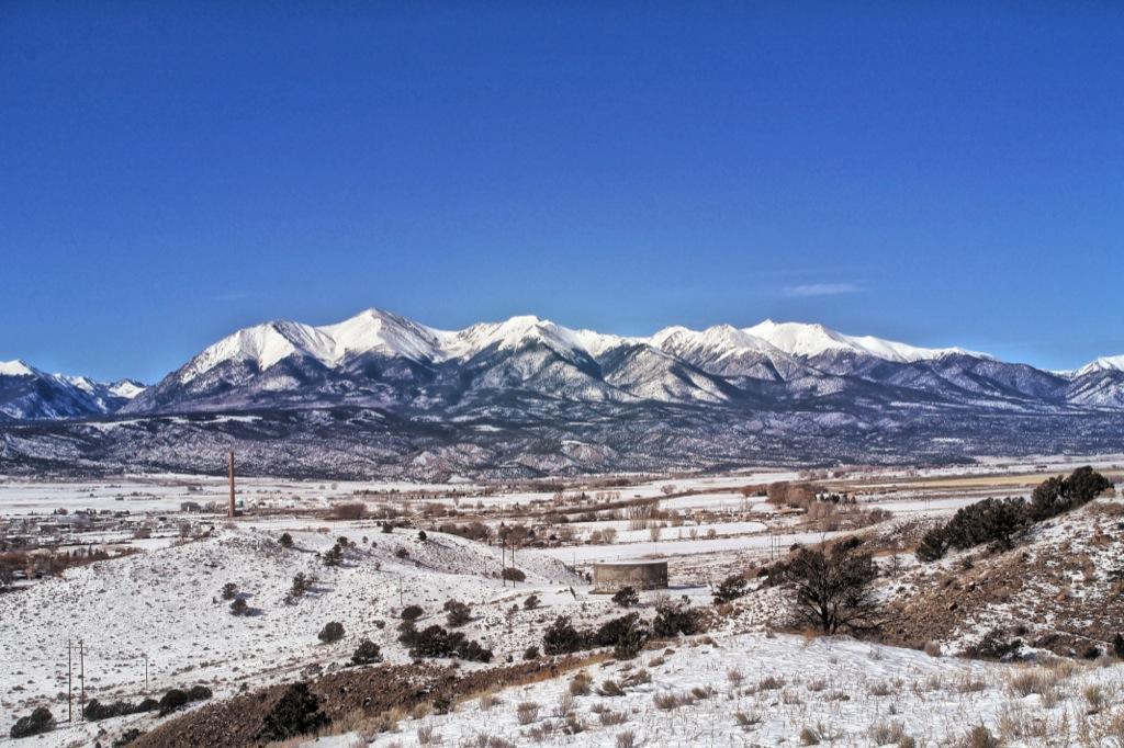 Snow-covered mountain range under a clear blue sky, with foreground featuring rolling hills and sparse vegetation. The landscape includes patches of snow and a distant view of a valley and structures. Arkansas Hills mountain bike trail.
