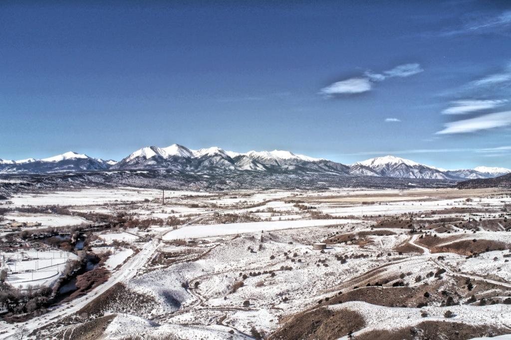 A panoramic view of snow-covered mountains under a clear blue sky, with a valley and winding river below. The landscape features rolling hills, patches of trees, and signs of winter's pristine beauty. Arkansas Hills mountain bike trail.