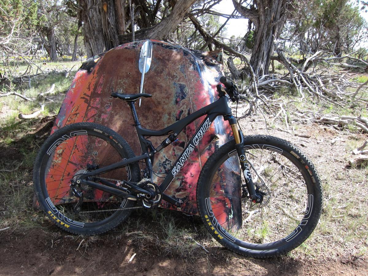 Santa Cruz Tallboy Carbon: A mountain bike leaned against a colorful, rusted metal structure in a forested area. The bike has large wheels and is marked with the brand name "Santa Cruz," surrounded by trees and underbrush.