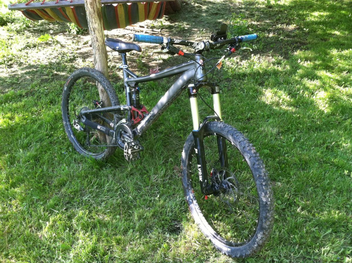 Diamondback Mission Pro: A mountain bike resting on green grass, featuring a silver frame, thick tires, and blue handlebar grips. The bike has a dual suspension system, with visible disc brakes and a sturdy front fork. In the background, there is a colorful hammock hanging from a tree.