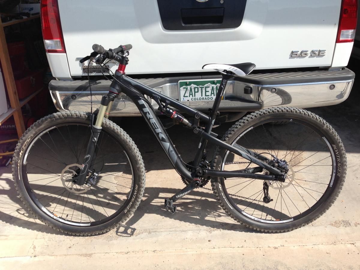 Gary Fisher Rumblefish: A black Trek mountain bike positioned next to the rear of a white vehicle, with the vehicle's license plate visible. The bike features thick tires, a suspension fork, and a sleek design. The background includes some garage clutter, indicating a casual outdoor setting.