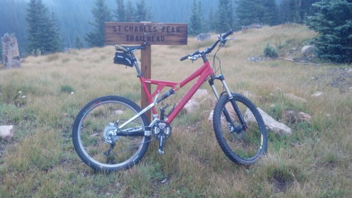 Yeti 575: A mountain bike leaning against a wooden trailhead sign that reads "St. Charles Peak Trailhead." The bike is red with black accents and positioned on grassy terrain surrounded by trees in a misty, mountainous landscape.