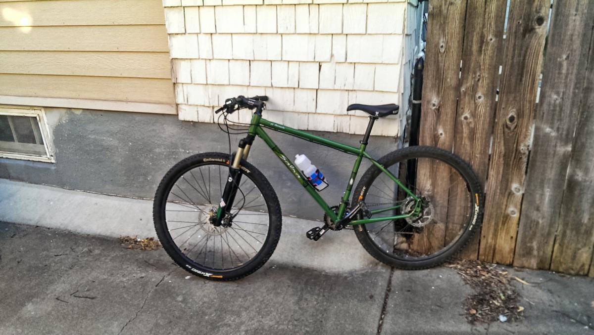 Salsa El Mariachi 3: A green mountain bike parked next to a wooden fence and a house with light-colored siding. The bike features thick tires, a water bottle holder, and is positioned against a textured wall.