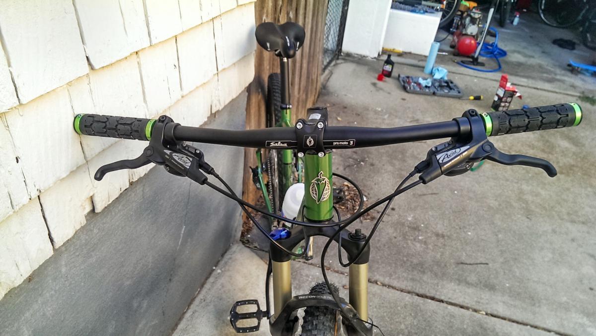 Salsa El Mariachi 3: Alt text: A close-up view of a mountain bike's handlebars, featuring black grips with green accents, brake levers, and a stem marked with the brand "Salsa." In the background, a green bicycle and a garage environment are visible.