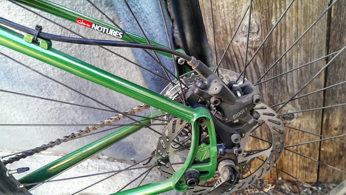 Salsa El Mariachi 3: Close-up view of a bicycle's rear brake system, showcasing a green bicycle frame, disc brake rotor, and caliper. The image highlights the detailed components including the chain, brake line, and a wooden background. Suitable for discussions on mountain biking or bicycle maintenance.