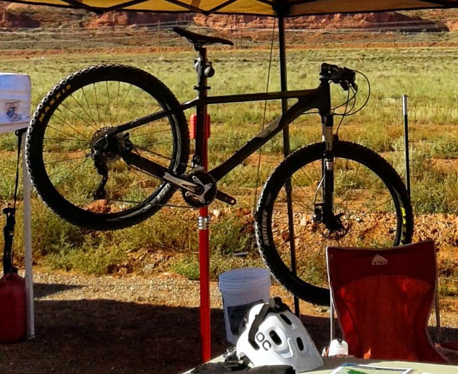 Foundry Cycles Tomahawk: A mountain bike is suspended on a repair stand beneath a canopy, with a grassy area and red rock formations in the background. A folding chair and a cycling helmet are visible in the foreground.