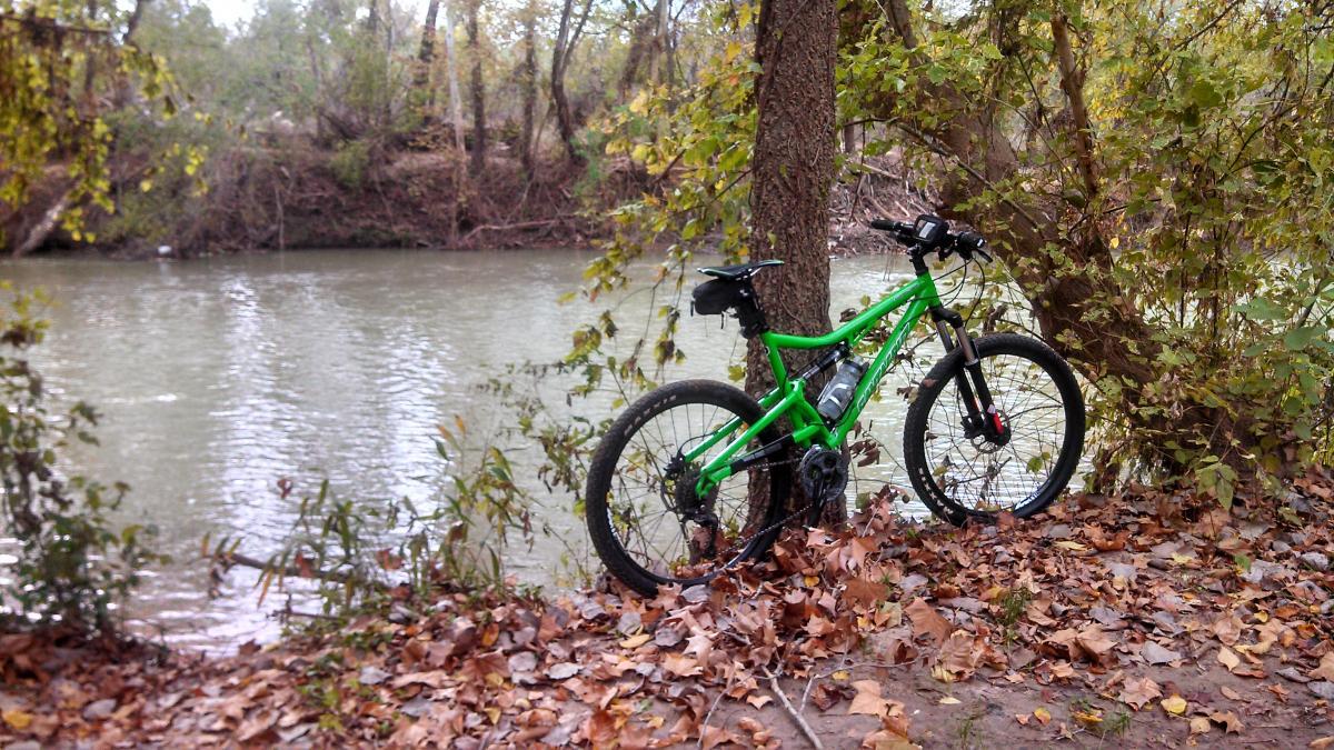 A green mountain bike parked next to a river, surrounded by autumn leaves and trees. The water reflects the surrounding landscape, creating a tranquil outdoor scene. Colorado River Refuge mountain bike trail.