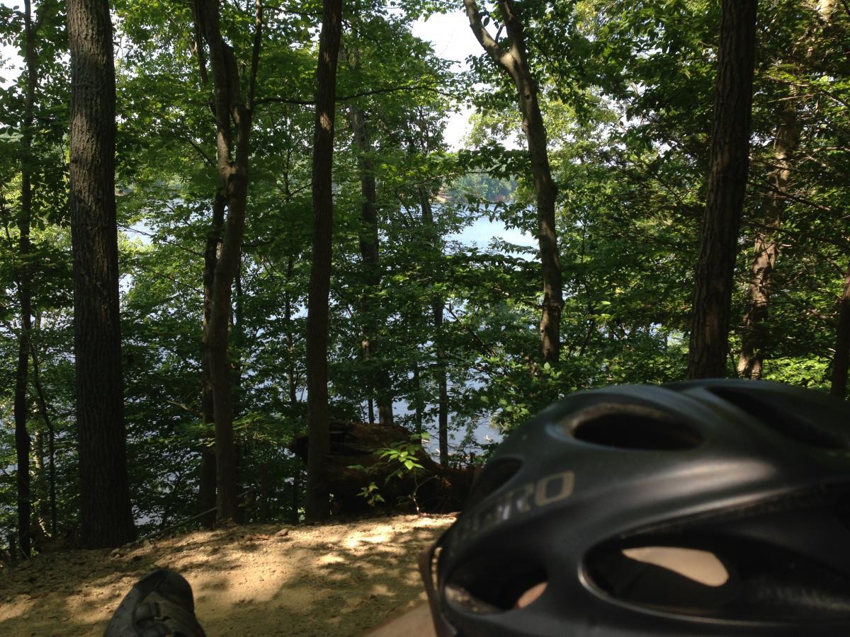 A view of a tranquil forest scene overlooking a lake, featuring tall trees with lush green leaves. In the foreground, a black bicycle helmet rests on sandy ground, suggesting a nearby biking or outdoor activity. Sunlight filters through the trees, creating a peaceful atmosphere. Fountainhead Regional Park mountain bike trail.