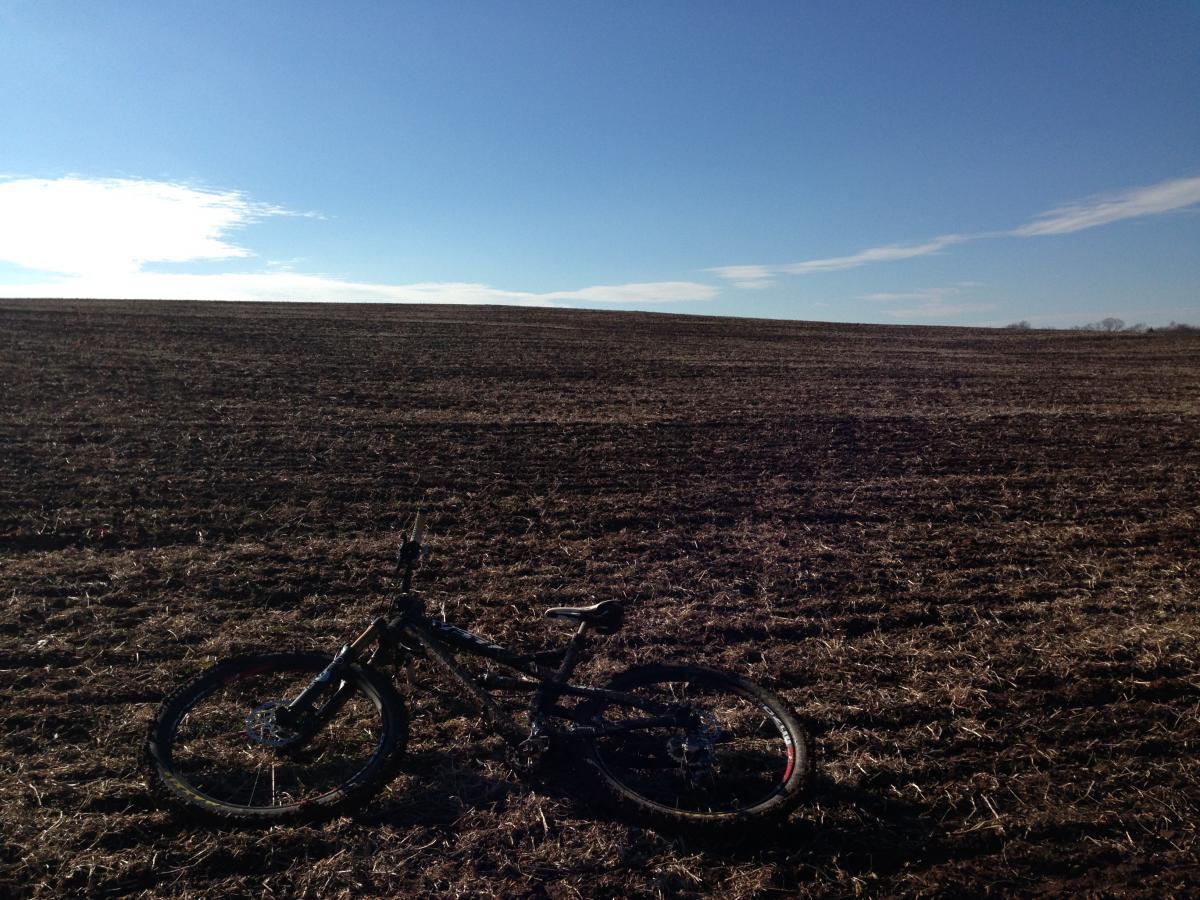 A mountain bike resting on the ground of a plowed field under a clear blue sky, with a gentle horizon in the background. Schaeffer Farms mountain bike trail.