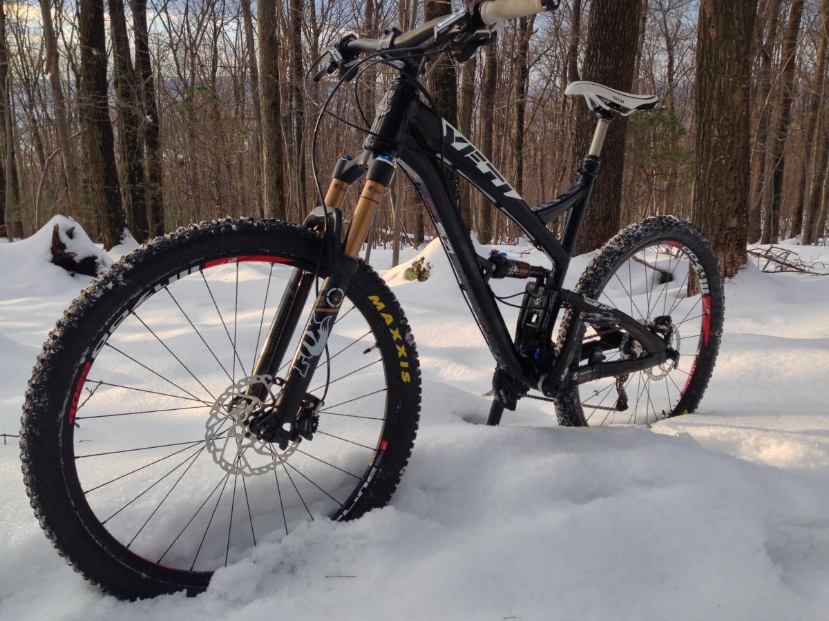 Yeti SB-75: A mountain bike resting on a snowy forest trail, surrounded by trees. The bike has a black frame with yellow and red accents, and its tires are equipped for snow conditions. Sunlight filters through the trees, illuminating the scene.