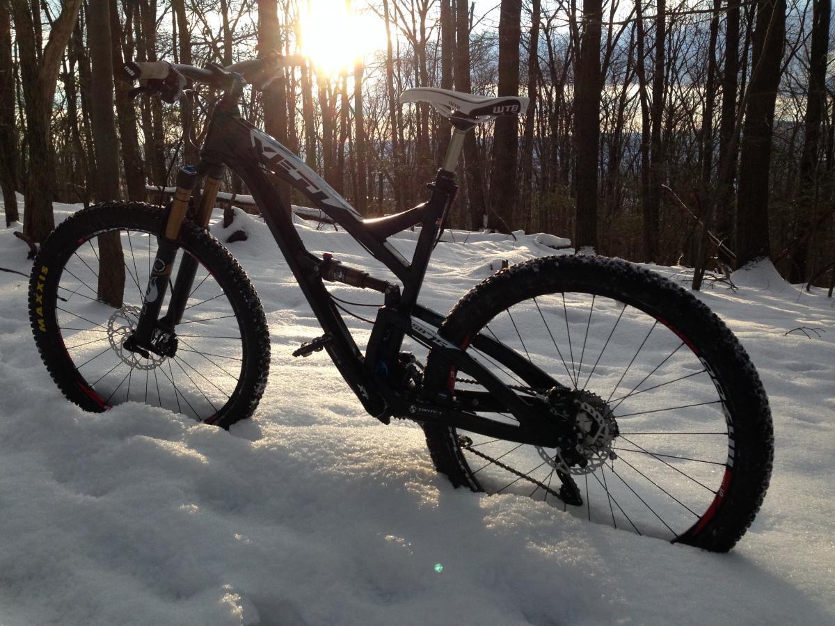 A mountain bike stands in a snowy forest, with trees and a soft sunlight filtering through the branches in the background. The bike features large tires suitable for winter riding, and the snow is freshly fallen around it. Gambrill State Park mountain bike trail.