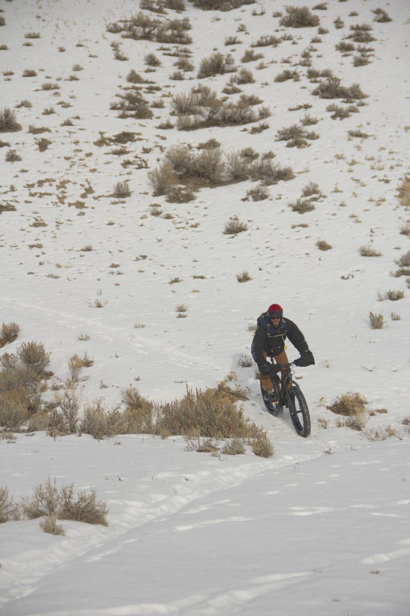 A person riding a fat bike on a snowy landscape, with patches of grass and shrubs visible in the background. The scene captures a winter biking adventure, highlighting the white snow and rugged terrain. Discovery Hill Trails mountain bike trail.