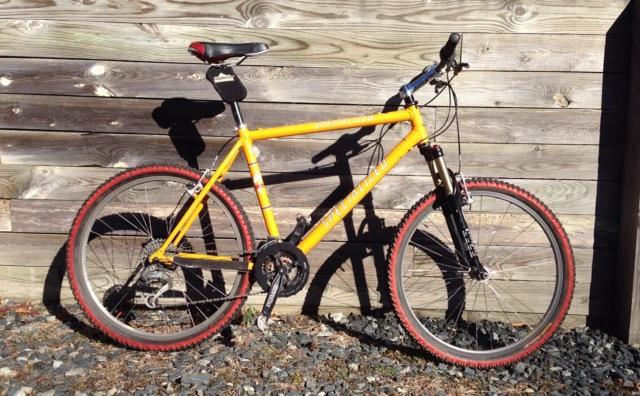 Specialized S-Works: A bright yellow mountain bike with red tires, resting against a wooden wall. The bike features a black seat, front suspension, and multiple gears, with a gravel surface beneath it.