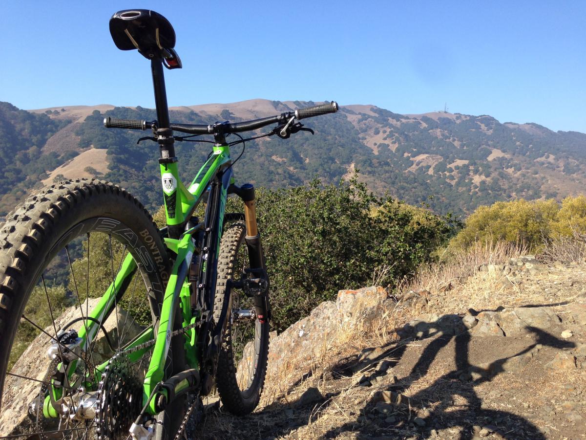 Trek Slash: A vibrant green mountain bike resting on a rocky outcrop, with a panoramic view of rolling hills and blue skies in the background. The bike's rear wheel is prominently featured, highlighting its rugged tires, while the lush vegetation and distant mountains create a scenic outdoor setting.