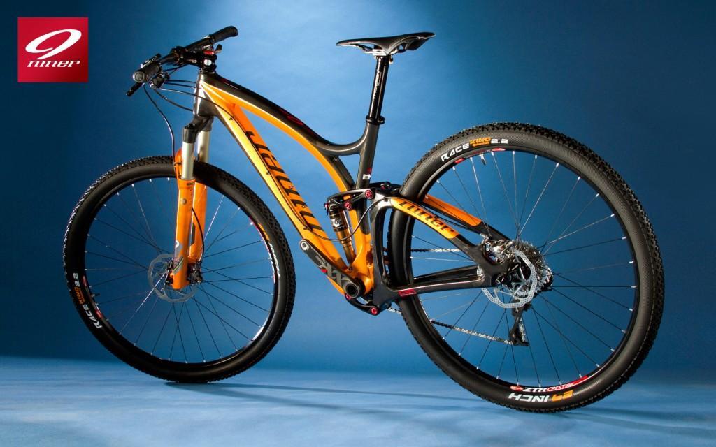 Niner Jet 9 RDO: High-performance mountain bike with an orange and black design, displayed against a blue background. The bike features large wheels and a lightweight frame, suitable for off-road cycling. The logo of the brand "Niner" is visible in the top left corner.