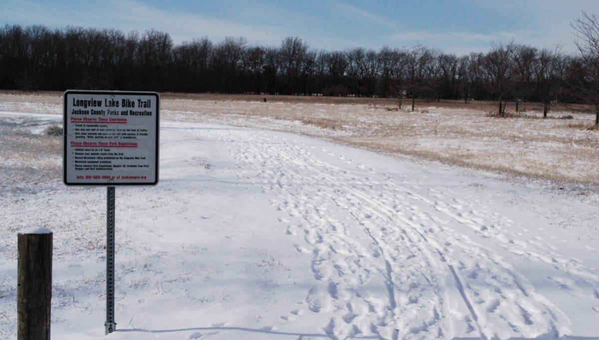 A snow-covered bike trail entrance featuring a sign for the Longview Lake Bike Trail, located in Jackson County Parks and Recreation. The sign provides information and guidelines for park visitors, with a backdrop of a field and sparse trees in winter. Longview Lake mountain bike trail.