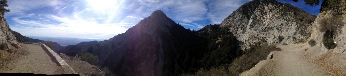 A panoramic view of a mountainous landscape, showcasing rocky peaks and rugged terrain under a partly cloudy sky. A dirt path leads along the edge of a cliff, with dense evergreen trees visible in the lower areas. The scene captures the beauty of nature, highlighting the elevation and expansive views of the valley below. Mount Lowe Railway mountain bike trail.