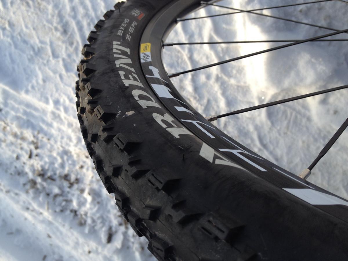 Maxxis Ardent: Close-up image of a bike tire with a rugged tread pattern, resting on snow. The tire features prominent markings and is partially angled to show the sidewall and wheel spokes in the background.