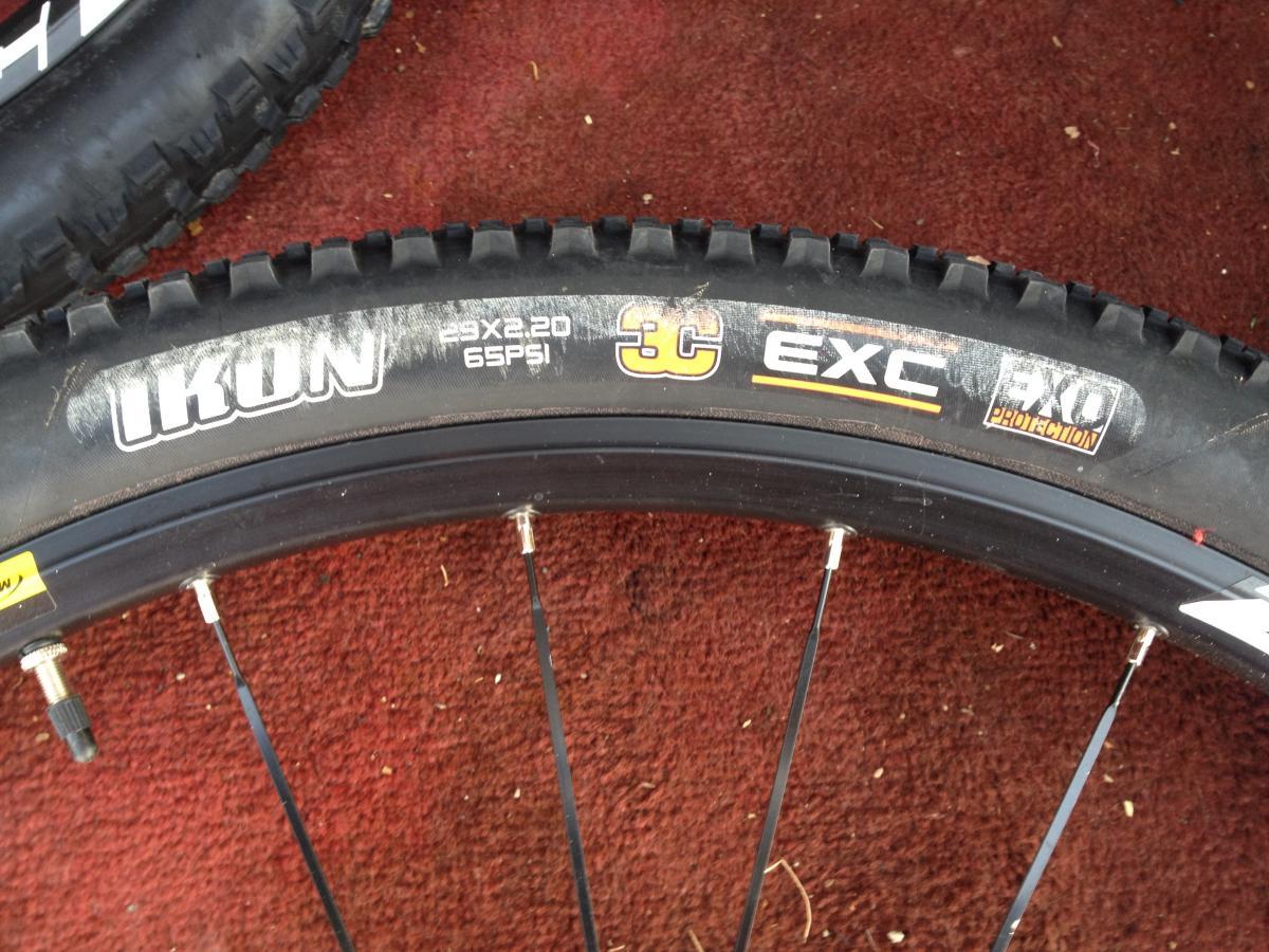 Maxxis Ikon: Close-up view of a mountain bike tire, featuring the brand logo "Ikon" and specifications indicating a size of 29 x 2.20 inches with a pressure rating of 65 PSI. The tire includes markings for "3C EXC" and "PRO Protection," indicating advanced features for durability and performance. The background shows a red textured surface.