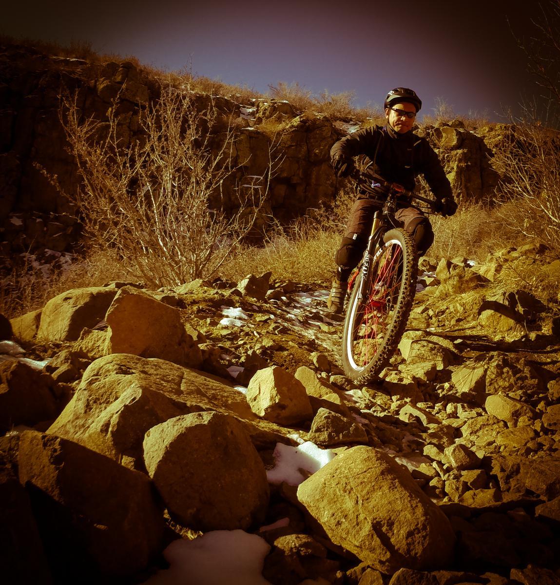 A person riding a mountain bike on a rocky trail, surrounded by sparse vegetation and a rocky landscape. The scene is illuminated by warm lighting, suggesting a late afternoon or early evening. North Table Mountain mountain bike trail.